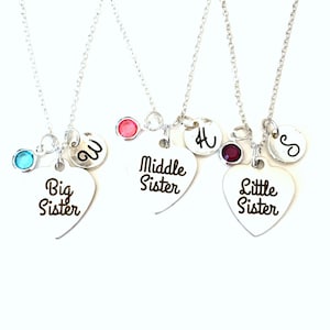 May include: Three silver necklaces with heart-shaped pendants. Each pendant has the words "Big Sister", "Middle Sister", and "Little Sister" printed on them. Each necklace has a different colored gem and a letter charm.
