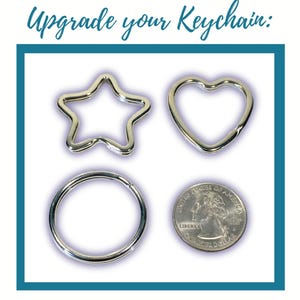 Puede incluir: Un conjunto de anillas para llaveros de metal plateado con formas de estrella, corazón y círculo, junto a una moneda para comparar el tamaño. El texto "Upgrade your Keychain:" aparece impreso en azul sobre los anillos.