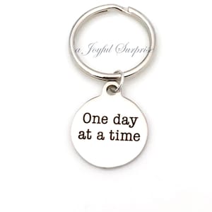 Può includere: Portachiavi d'argento con un'etichetta rotonda che dice "One day at a time".