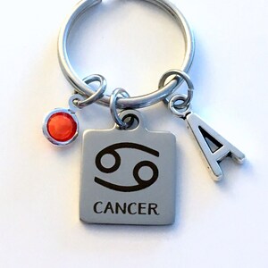 May include: A silver keychain with a red gem, a silver charm with the astrological symbol for Cancer, and a silver charm with the letter A. The charm with the Cancer symbol also has the word "Cancer" engraved on it.