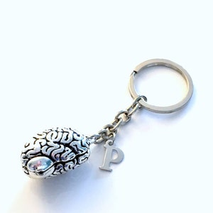 Può includere: Portachiavi in ​​argento con un ciondolo a forma di cervello dettagliato e un ciondolo a forma di lettera 'P'. Il portachiavi è attaccato a un anello per chiavi in ​​argento.