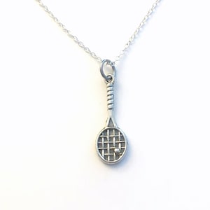 Op de afbeelding: Een zilverkleurige tennisracket hanger met een kleine bal hangt aan een fijne zilveren ketting. Het racket heeft een gedetailleerde handgreep en een netachtig ontwerp. De ketting wordt gepresenteerd tegen een witte achtergrond.