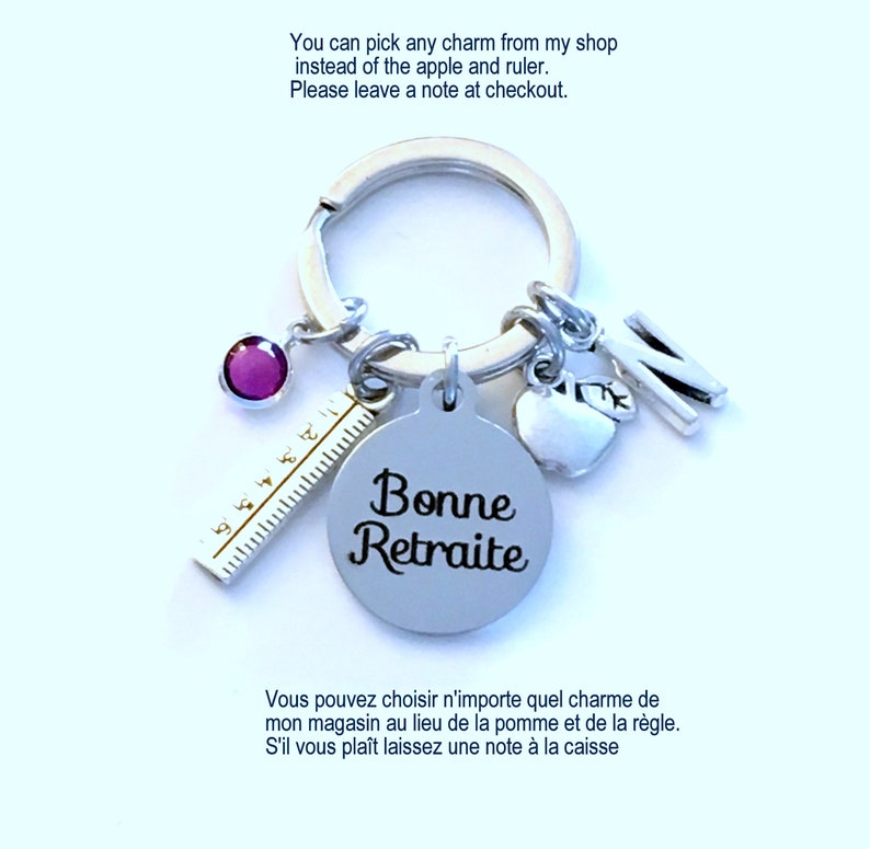 May include: Silver keychain with multiple charms, including a purple gem, a ruler, a round tag with "Bonne Retraite" text, an apple, and the letter "N". The keychain also has text: "You can pick any charm from my shop instead of the apple and ruler."