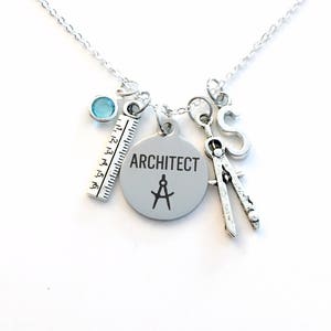 Könnte beinhalten: Silberkette mit Anhängern zum Thema Architekt. Die Kette hat ein Lineal, einen Zirkel, den Buchstaben "S", einen runden Anhänger mit dem Wort "ARCHITECT" und einem Kompasssymbol sowie einen kleinen blauen Edelstein. Die Anhänger hängen an einer filigranen Kette.