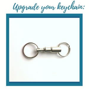 Op de afbeelding: Een zilverkleurige metalen sleutelhangerconnector met snelsluiting, voorzien van een cilindrisch middenmechanisme met een verend ontwerp en twee ronde splitringen aan elk uiteinde. De tekst Upgrade your keychain is zichtbaar.