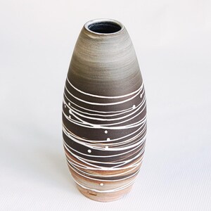 Könnte beinhalten: Eine handgefertigte Keramikvase mit einem einzigartigen Design. Die Vase hat einen dunkelbraunen Sockel, der sich nach oben hin zu hellbraun und grau verjüngt. Weiße, wellenförmige Linien und Punkte umschließen die Vase und bilden ein dekoratives Muster. Die Vase hat eine glatte, glänzende Oberfläche.