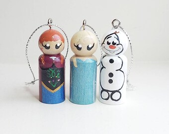 Può includere: Tre bambole in legno dipinte a mano con cordini argentati per appenderle. Le figure presentano abiti distinti: una con capelli rossi e vestito viola e blu, una con capelli biondi e vestito azzurro, e un pupazzo di neve bianco con bottoni.