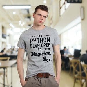 Op de afbeelding: Een grijs T-shirt met zwarte tekst die "Ik ben een Python-ontwikkelaar Net als een tovenaar" luidt, met een zwart-witte illustratie van een hoge hoed en een toverstaf.