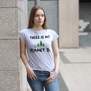 May include: A woman wearing a grey t-shirt with the text "There is no Planet B" and a graphic of three trees. She is standing in front of a concrete wall.