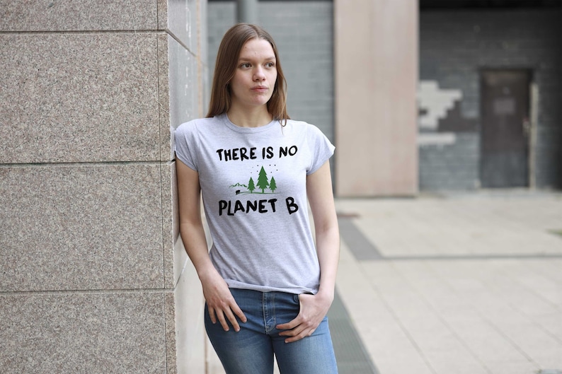 May include: A woman wearing a grey t-shirt with the text "There is no Planet B" and a graphic of three trees. She is standing in front of a concrete wall.