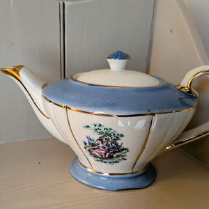 Op de afbeelding: Crèmekleurige porseleinen theepot met een lichtblauwe band en gouden details. De theepot heeft een decoratieve scène van twee figuren onder een boom. De deksel en voet zijn ook lichtblauw. Een klassiek stuk voor theeliefhebbers.