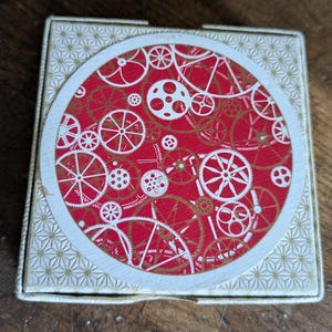 May include: A square box with a lid featuring a circular center design. The center displays a red background with a repeating pattern of white and gold mechanical gears and cogs. The border of the lid has a white and gold geometric star pattern.