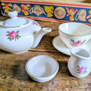 Puede incluir: Un juego de té de porcelana blanca con motivos de rosas rosas y bordes dorados. El conjunto incluye una tetera con tapa, una taza de té sobre su plato, una pequeña jarra de leche y un cuenco poco profundo, presentados con una caja decorativa.