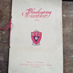 May include: Vintage Thanksgiving program from 1942. The cover is cream-colored with red text and a decorative floral design. The program features the RTC logo and the text "Battery F, 32nd Battalion, Field Artillery Replacement Training Center, Fort Sill, Oklahoma."