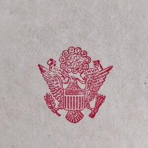 May include: A red ink stamp of the Great Seal of the United States. The design features a bald eagle with outstretched wings, holding arrows and an olive branch. Above the eagle is a banner with the words "E PLURIBUS UNUM".