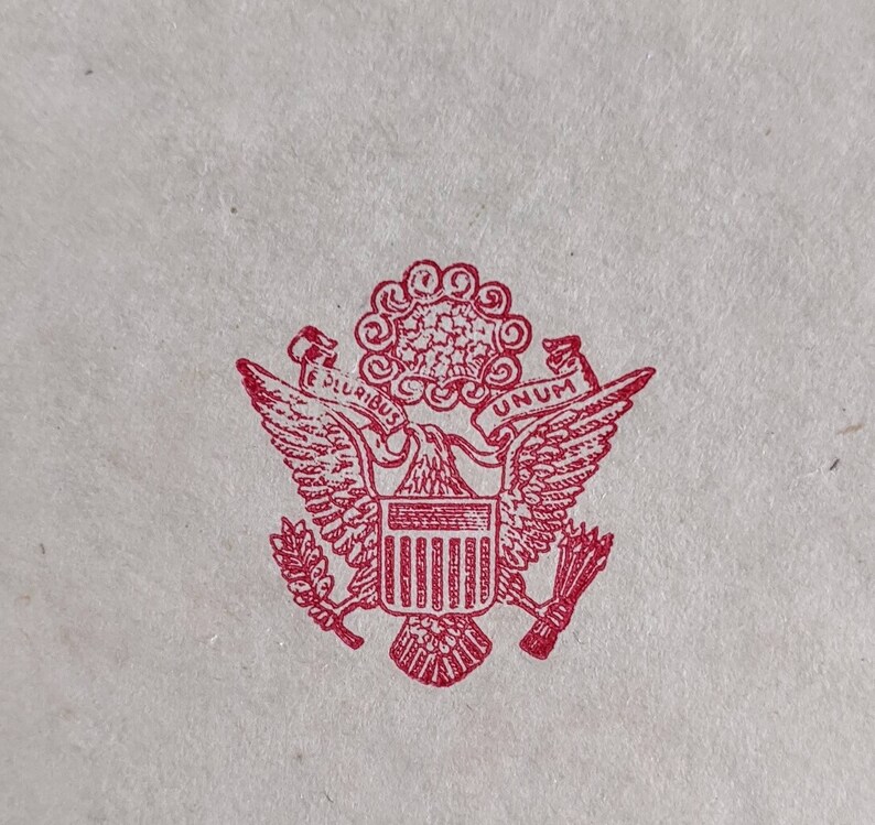 May include: A red ink stamp of the Great Seal of the United States. The design features a bald eagle with outstretched wings, holding arrows and an olive branch. Above the eagle is a banner with the words "E PLURIBUS UNUM".