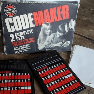 May include: A vintage Airfix Codemaker game box with two black plastic code wheels. The box features a red and white logo and the text "Codemaker" in large red letters. The box also includes the text "2 Complete Sets, One for you and one for a friend. Send and receive coded messages. Full instructions on back of box."