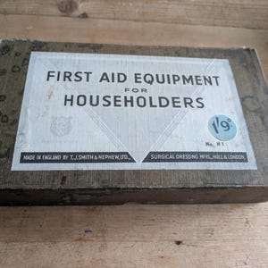 May include: A vintage wooden first aid kit with a white label. The label reads "FIRST AID EQUIPMENT FOR HOUSEHOLDERS" and includes the text "MADE IN ENGLAND BY T.J. SMITH & NEPHEW, LTD." and "SURGICAL DRESSING MFRS, HULL & LONDON."