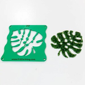 Peut inclure: Un pochoir en plastique vert et une feuille décorative verte. Le pochoir présente un motif de feuille de monstera et le texte "www.FritForming.com". La feuille décorative est verte, brillante et texturée.