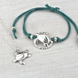 May include: A silver turtle necklace and a teal cord bracelet with a silver turtle charm. Both pieces of jewelry feature a detailed turtle design.