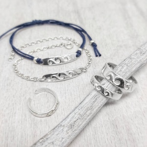 Può includere: Un set di gioielli in argento con un motivo a onde, che comprende due braccialetti, uno con un cordoncino blu e uno con una catena d'argento, e due anelli per le dita dei piedi.