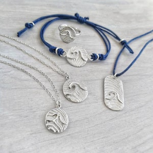 Könnte beinhalten: Ein Set aus silbernem Wellenschmuck, bestehend aus einem Ring, einem Armband und zwei Halsketten. Der Schmuck hat ein strukturiertes Wellendesign und wird auf einer hellbraunen Holzoberfläche präsentiert.
