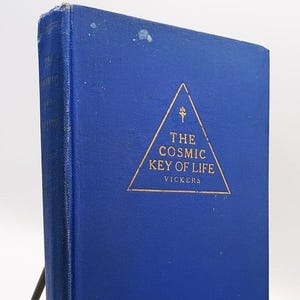 May include: A vintage book titled "The Cosmic Key of Life" by Vickers. The book has a blue cover with gold lettering and a geometric design. The book is displayed on a black metal stand.