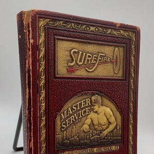May include: A vintage, burgundy-colored book with gold embossed lettering and designs. The cover features the words "SURE FIRE" and "MASTER SERVICE INC." with an illustration of a man. The book is displayed on a black stand.