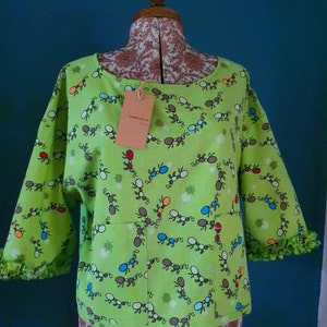 May include: A lime green blouse with a pattern of colourful ants. The blouse has a scoop neck and 3/4 sleeves with ruffles at the cuffs. The blouse has a pocket on the front with the text "The Doodle" printed on a tag.
