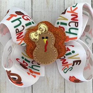 May include: A white ribbon bow with the text "Give Thanks" printed on it. The bow has a felt turkey applique with a gold bow on top.