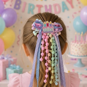 May include: A hair bow featuring a felt birthday cake applique with three candles, layered with blue gingham ribbon, and multiple strands of pastel pink, yellow, and blue pom-pom trim that hang down to create a festive, multi-textured hair accessory.
