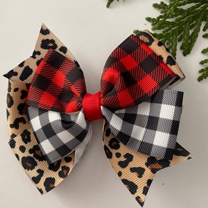 May include: A layered hair bow featuring a mix of patterns. The bow has a red center and is composed of ribbons with leopard print, red and black buffalo plaid, and black and white gingham patterns. A sprig of greenery is in the background.