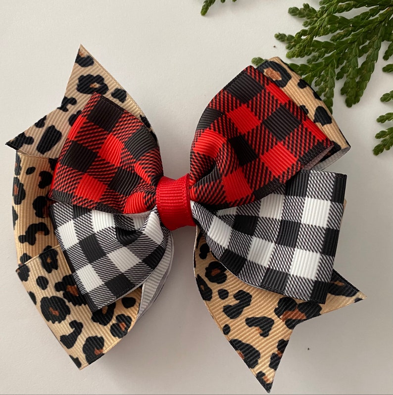 May include: A layered hair bow featuring a mix of patterns. The bow has a red center and is composed of ribbons with leopard print, red and black buffalo plaid, and black and white gingham patterns. A sprig of greenery is in the background.
