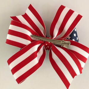 May include: Red, white, and blue striped ribbon bow with a blue and white star patterned ribbon in the center. The bow is attached to a silver alligator clip.