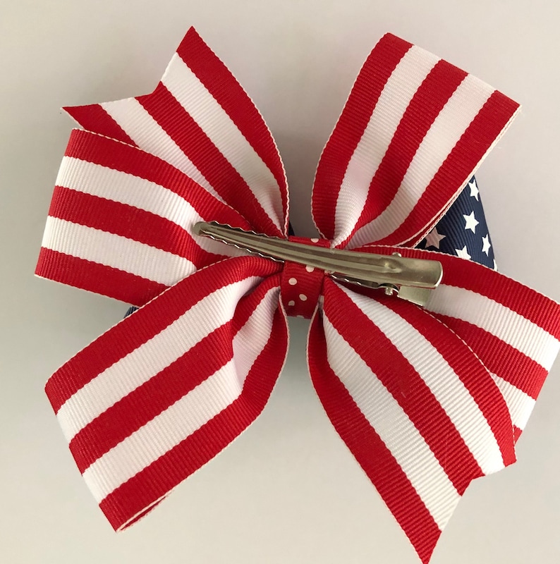 May include: Red, white, and blue striped ribbon bow with a blue and white star patterned ribbon in the center. The bow is attached to a silver alligator clip.