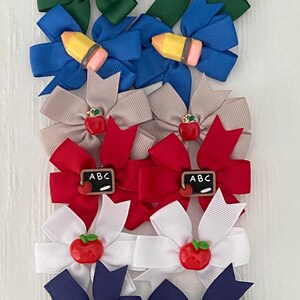 May include: A collection of colorful hair bows in various shades of blue, green, red, white, and tan. Some bows feature apple or pencil embellishments, while others have small chalkboard accents with the letters "ABC".