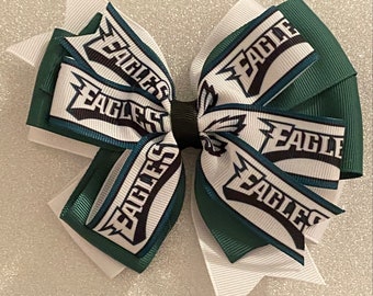 May include: A handmade hair bow featuring the word "EAGLES" in white, black, and teal. The bow is made of layered ribbons in white, dark green, and teal. The bow is tied with a black center.