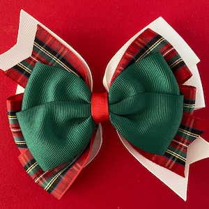 May include: A layered hair bow with a festive Christmas theme. The bow features a combination of white, green, red, and plaid ribbons. The center is a solid green, with a red band in the middle. The background is a solid red.