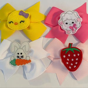 May include: Four Easter-themed hair bows with different embroidered designs: a yellow chick, a white lamb, a white bunny with a carrot, and a red strawberry. The bows are made of yellow, pink, white, and light pink ribbon.