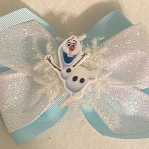 May include: A blue and white glitter bow with a white Olaf character from the movie Frozen. The bow is decorated with white glitter and a snowflake design.