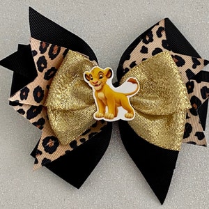 May include: A black, gold, and brown bow with a lion king character in the center. The bow is made of ribbon and has a leopard print pattern.