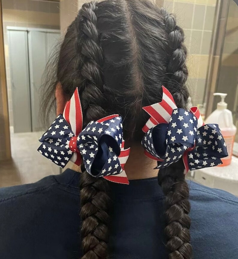 May include: Two braided hairstyles with red, white, and blue star-patterned hair bows.