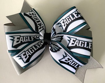 May include: A grey and white hair bow with a green and black ribbon featuring the words "Eagles" repeated. The bow is tied in a large, symmetrical shape.