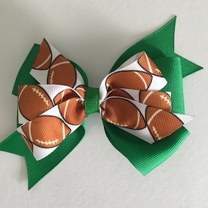 May include: A green and white hair bow with a football pattern. The bow is made of two layers of ribbon, with the top layer being white with brown footballs and the bottom layer being solid green.