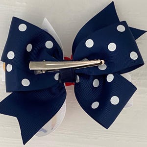 Op de afbeelding: Een navyblauwe haarstrik met witte stippen en een zilveren alligatorclip. De strik is in een klassieke stijl geknoopt met een wit midden.