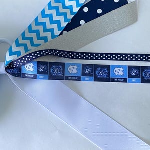 May include: A set of hair ties featuring a variety of ribbons in blue, white, and silver. The ribbons have different patterns, including polka dots, chevron, and a repeating pattern of the University of North Carolina Tar Heels logo. The text "Tar Heels" is visible on the ribbon.