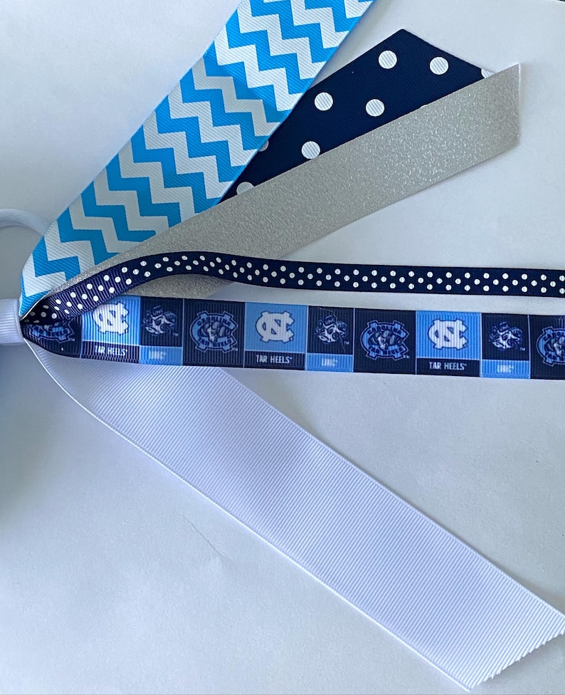 May include: A set of hair ties featuring a variety of ribbons in blue, white, and silver. The ribbons have different patterns, including polka dots, chevron, and a repeating pattern of the University of North Carolina Tar Heels logo. The text "Tar Heels" is visible on the ribbon.