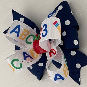 Op de afbeelding: Een wit en marineblauw haarband met witte stippen. De strik heeft een wit lint met de letters ABC en het getal 3 erop gedrukt. In het midden van de strik zit een rode appelbedel.