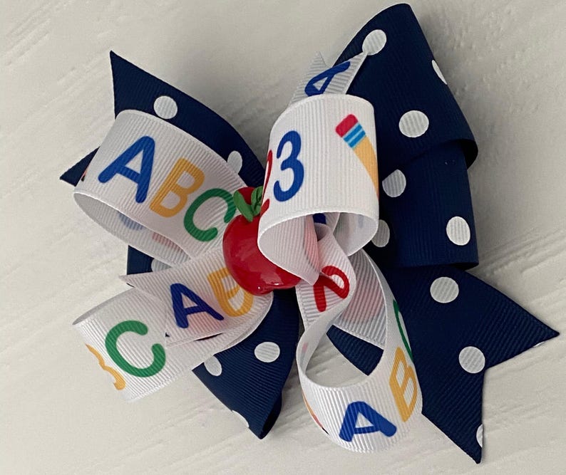 Op de afbeelding: Een wit en marineblauw haarband met witte stippen. De strik heeft een wit lint met de letters ABC en het getal 3 erop gedrukt. In het midden van de strik zit een rode appelbedel.
