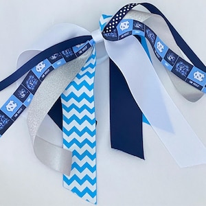 May include: A blue and white hair tie with a chevron pattern, a silver glitter ribbon, a white ribbon, a navy blue ribbon, and a ribbon with the UNC Tar Heels logo and the words "Tar Heels".